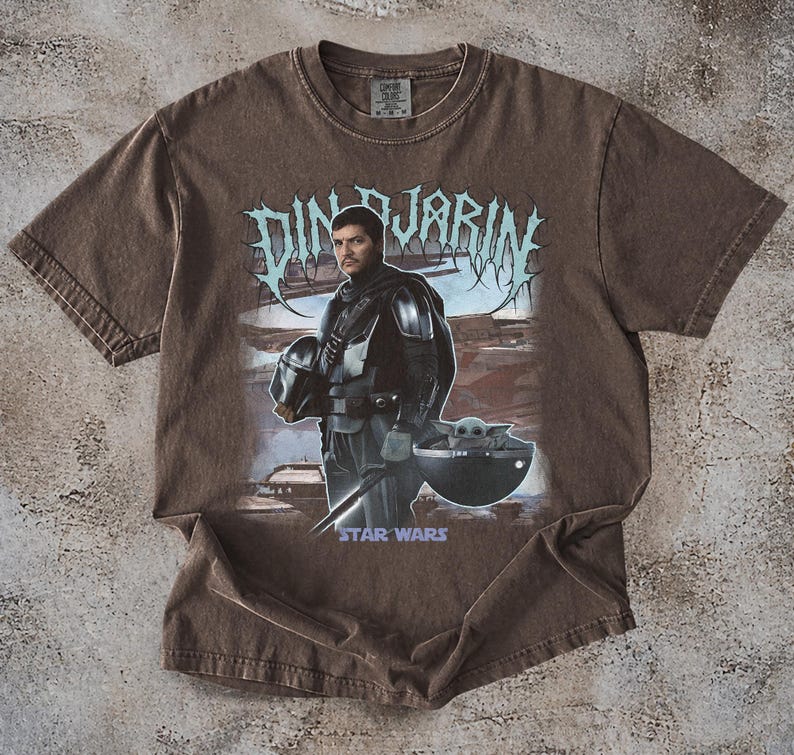 May include: Brown t-shirt featuring a graphic of Din Djarin from Star Wars, holding his helmet, with Grogu in a floating pod. The text "DIN DJARIN" is above the image, and "STAR WARS" is below.