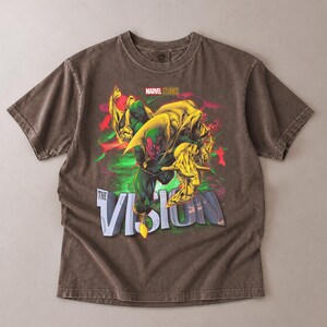 Puede incluir: Una camiseta marr&oacute;n con un gr&aacute;fico del personaje de Marvel, Vision. El dise&ntilde;o incluye a Vision en verde y amarillo, con el texto "THE VISION" en blanco y azul. El logotipo de Marvel Studios est&aacute; en la parte superior.