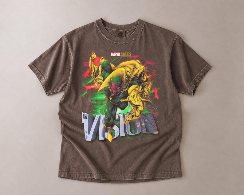 Puede incluir: Una camiseta marr&oacute;n con un gr&aacute;fico del personaje de Marvel, Vision. El dise&ntilde;o incluye a Vision en verde y amarillo, con el texto "THE VISION" en blanco y azul. El logotipo de Marvel Studios est&aacute; en la parte superior.