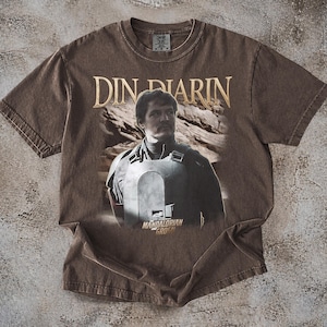 May include: Brown t-shirt featuring a graphic of Din Djarin from The Mandalorian. The text "Din Djarin" is printed in gold above the image. The words "Mandalorian Grogu" are printed below the image.