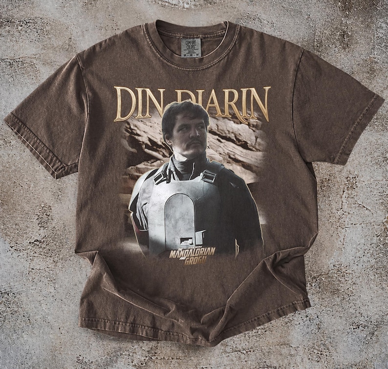 May include: Brown t-shirt featuring a graphic of Din Djarin from The Mandalorian. The text "Din Djarin" is printed in gold above the image. The words "Mandalorian Grogu" are printed below the image.