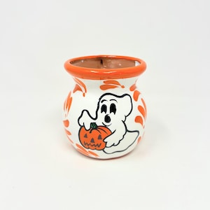 May include: A small, round ceramic pot with a white base and a bright orange rim. The exterior features a hand-painted illustration of a white ghost holding a carved orange jack-o'-lantern pumpkin, surrounded by orange leaf-like decorative accents.