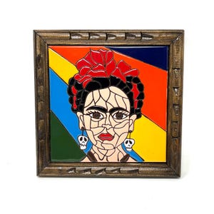 May include: A framed ceramic tile featuring a colorful portrait of a woman with a red flower in her hair and skull earrings. The background is a vibrant diagonal pattern of red, orange, yellow, blue, and green. The frame is a dark brown color.