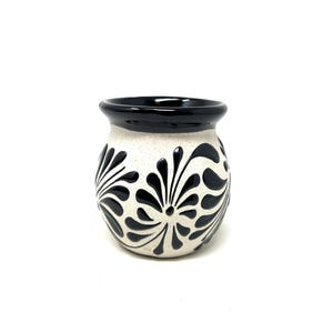 May include: A small, round ceramic vase with a speckled cream-colored body. The vase features a black rim and is decorated with a repeating black floral pattern. The design is symmetrical and covers the majority of the vase's surface.