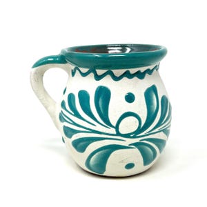 May include: A white ceramic mug with a teal handle and rim. The mug features a teal floral design and is suitable for hot or cold beverages. The mug is approximately 4 inches tall and 3 inches in diameter.