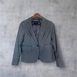 Puede incluir: Una blazer gris con cierre de un solo botón y dos bolsillos delanteros cuelga de una percha de madera. La blazer tiene un corte entallado y un diseño clásico, adecuado para uso profesional o informal. El forro es de un gris más oscuro.
