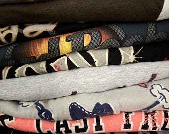 May include: A stack of folded t-shirts in various colors. The top shirt is gray with the word "DREAM" in brown lettering. Other shirts feature graphic designs, including a snake, a cartoon alligator, and text.