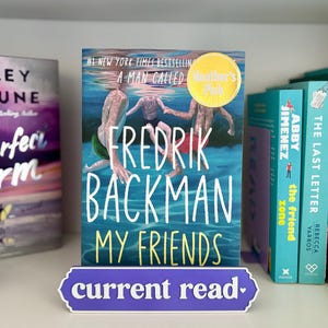 May include: A book titled My Friends by Fredrik Backman with a cover illustration of three people swimming underwater. The book is displayed behind a purple sign that reads current read. A yellow circular sticker on the cover says Heather's Pick.