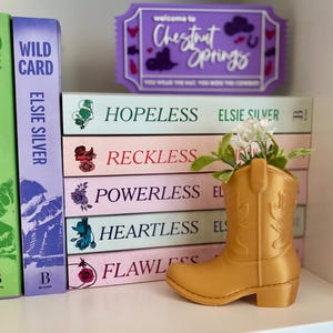 May include: A gold-coloured cowboy boot-shaped planter containing small white flowers, positioned in front of a stack of five books titled Hopeless, Reckless, Powerless, Heartless, and Flawless by Elsie Silver, with a purple Chestnut Springs sign behind.