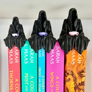 May include: Three black hooded figures in varying sizes perched on top of five books by Sarah J. Maas. Each figure wears a dark cloak and holds a small, solid-coloured teacup in shades of light purple, lavender, and pale pink in their hands.