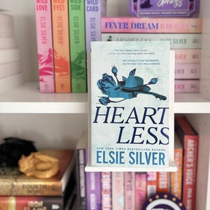 May include: A book titled Heartless by Elsie Silver displayed on a white shelf. The cover features a blue illustration of a cowboy hat, guitar, and roses. Surrounding the book are various other novels with colourful spines and a purple sign.