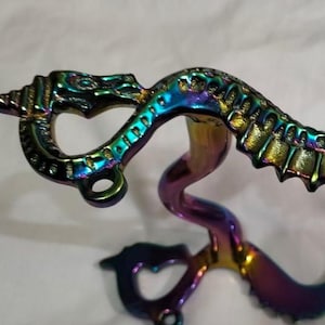 Puede incluir: Un bocado de caballo con diseño de dragón. El bocado es de metal y presenta un acabado iridiscente arcoíris. La cabeza del dragón está en la parte superior, y el bocado se curva hacia abajo con un anillo en cada extremo.