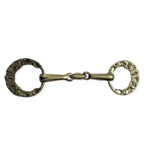 May include: A gold-colored horse bit with ornate, floral-like detailing on the circular cheek pieces. The bit has a central joint and is designed for use in equestrian activities. The metal has a polished finish.