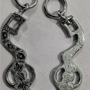 May include: Two silver-colored horse bits with floral and scrollwork designs. Each bit features a curved, decorative mouthpiece with a ring at the top and a loop at the bottom. The metal has a polished finish, reflecting light.