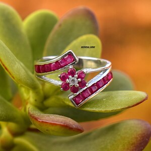 Può includere: Un anello d'argento con un motivo floreale. L'anello presenta un fiore composto da piccole pietre preziose rosse rotonde e un diamante centrale. Due fasce di pietre preziose rosse sono incastonate su entrambi i lati del fiore.