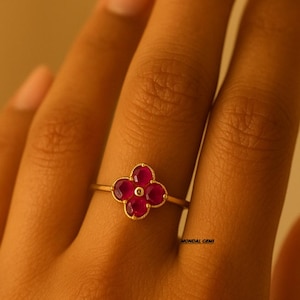 Peut inclure: Une bague en or avec un motif de fleur à quatre pétales. Chaque pétale est une pierre précieuse rouge vif, avec un petit accent doré au centre. La bague est présentée sur une main.