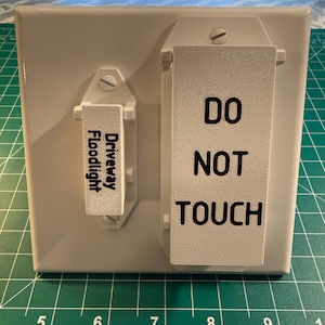 May include: A light gray electrical outlet cover with two attached labels. One label reads "Driveway Floodlight" and the other reads "DO NOT TOUCH" in black text. The cover is mounted on a green cutting mat.