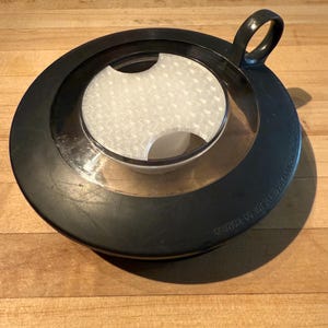 May include: A black lid with a clear center and a white filter. The lid has a handle and is sitting on a wooden surface. The text "CAUTION: DO NOT SEND HOT LIQUID" is printed on the lid.
