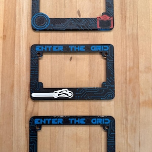 May include: Three black license plate frames with the text "ENTER THE GRID" in blue. The frames have a circuit board design and various colored accents, including red, yellow, and white. The frames are displayed on a wooden surface.
