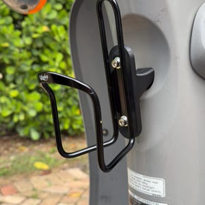 May include: Black metal water bottle holder mounted on a gray scooter. The holder is attached to the scooter with screws and has the word "Wake" printed on it. The scooter has a chrome mirror and orange turn signal.