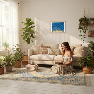 Könnte beinhalten: Ein gemütliches Wohnzimmer mit beigem Sofa, gemustertem Teppich und verschiedenen Topfpflanzen. Eine Frau sitzt auf dem Teppich und liest ein Buch. Ein gerahmtes Aquarell hängt an der Wand, und Sonnenlicht fällt durch ein Fenster.