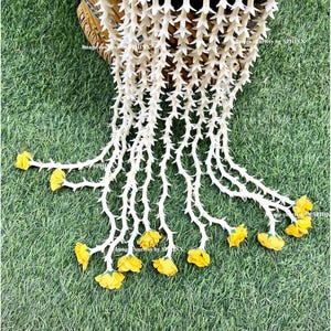 May include: A decorative garland with white, thorn-like elements cascading from a woven chair. The garland features yellow flower accents at the ends of the cascading strands, set against a backdrop of green grass.