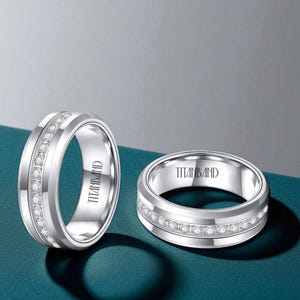 Puede incluir: Dos anillos plateados con una fila de pequeñas piedras transparentes en el centro. Los anillos tienen un acabado pulido y el texto "TITANBAND" está grabado en el interior. Los anillos se exhiben sobre una superficie turquesa.
