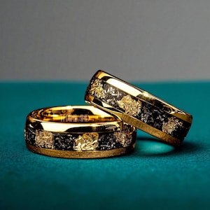 以下が含まれることがあります： 黒い象嵌と金箔のアクセントが付いた2つの金色の指輪。 リングは研磨仕上げで、バンドには「TITANBAND」の文字が見えます。 指輪はターコイズブルーの表面に表示されています。