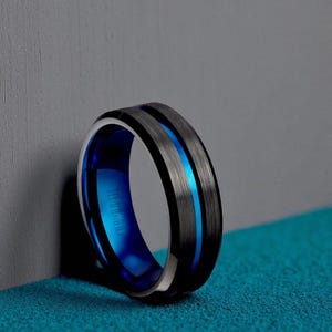 May include: A close-up of a black and silver ring with a bright blue interior and a blue stripe down the center. The ring is resting on a blue surface and leaning against a gray wall. The ring has the text "Titanium" on the inside.