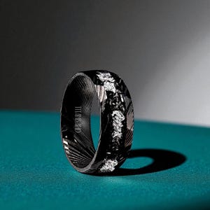 May include: A black ring with a textured pattern and silver accents. The ring has the text "TITAN BAND" on the side. The ring is set against a teal background.