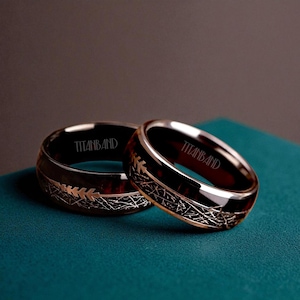 Könnte beinhalten: Zwei Ringe mit dunklem Holzmaserungs-Innenraum und silberfarbenem Metall-Außenbereich. Die Ringe haben ein dekoratives geätztes Design und das Wort "TITANBAND" ist auf der Innenseite des Rings eingraviert. Die Ringe werden auf einer türkisfarbenen Oberfläche präsentiert.