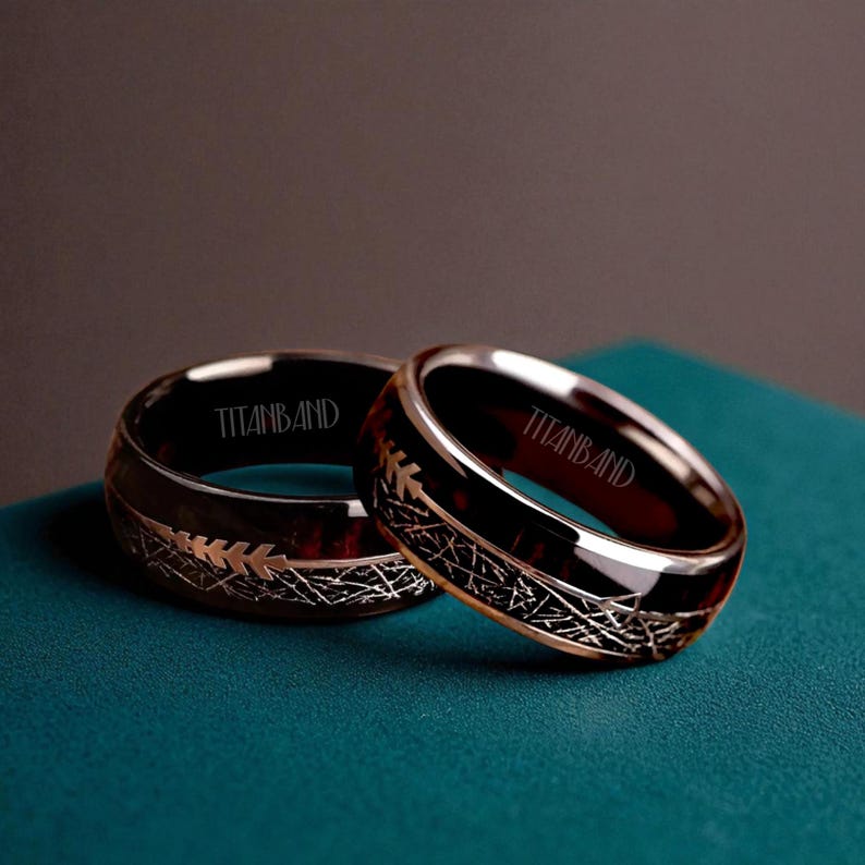 May include: Two rings with a dark wood-grain interior and silver-colored metal exterior. The rings have a decorative etched design and the word "TITANBAND" is engraved on the inside of the ring. The rings are displayed on a teal surface.