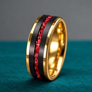 Op de afbeelding: Een ring met een gepolijste gouden band en een brede, geborstelde zwarte inleg in het midden. Een smal, getextureerd kanaal van felrode gemalen steen loopt horizontaal door het midden van de zwarte inleg voor een contrast.