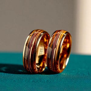 May include: Two gold wedding bands with a wood grain inlay and a polished finish. The rings have a band of thin, parallel lines and the text "TITANBAND" is visible on the side of one ring. The rings are displayed on a teal surface.