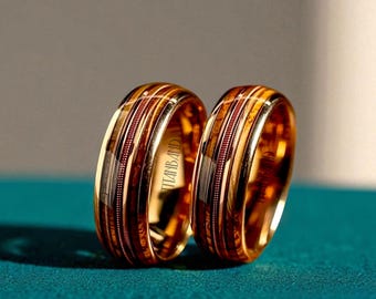 May include: Two gold wedding bands with a wood grain inlay and a polished finish. The rings have a band of thin, parallel lines and the text "TITANBAND" is visible on the side of one ring. The rings are displayed on a teal surface.