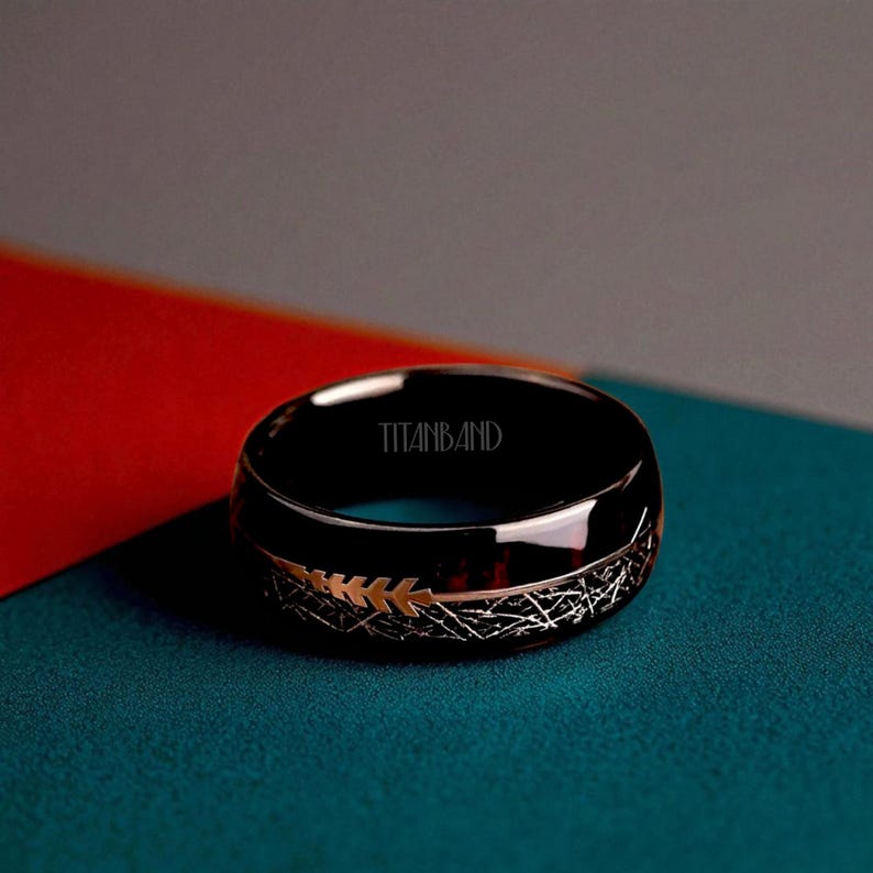 May include: A black ring with a gold and silver design. The ring has the text "TITANBAND" inside. The ring has a shiny finish and is set against a backdrop of red, gray, and teal.