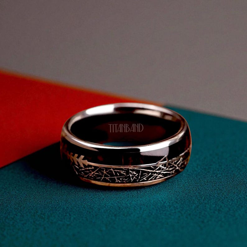 May include: A black and gold ring with the text "TITANBAND" engraved on the inside. The ring has a gold band with a black center and a decorative pattern. The ring is displayed on a teal surface with a red and gray background.
