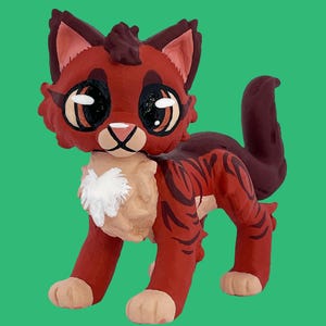 May include: A handcrafted, stylized cat figurine in shades of red, brown, and tan. The cat has large, expressive eyes, a white chest, and a striped pattern on its body. The figurine is posed on a green background.