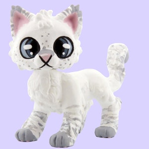 May include: A white figurine of a cat with gray markings and large blue eyes. The cat has pink inner ears and a gray nose. The figurine is standing on all fours with its tail curved upwards. The background is a light purple color.