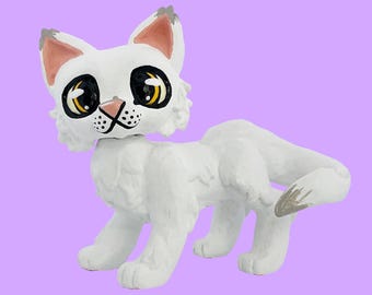 Könnte beinhalten: Eine weiße Katzenfigur mit großen, ausdrucksstarken Augen, einer rosa Nase und grauen Akzenten an Ohren und Schwanz. Die Katze ist in einer Laufhaltung vor einem lavendelfarbenen Hintergrund dargestellt. Die Figur hat ein strukturiertes, handbemaltes Aussehen.