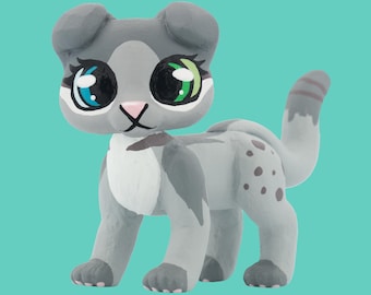 Könnte beinhalten: Handbemalte graue Katzenfigur mit großen, ausdrucksstarken Augen in Blau und Grün. Die Katze hat eine rosa Nase, eine weiße Brust und graue Flecken am Körper. Der Schwanz ist gestreift, und die Figur steht vor einem türkisfarbenen Hintergrund.