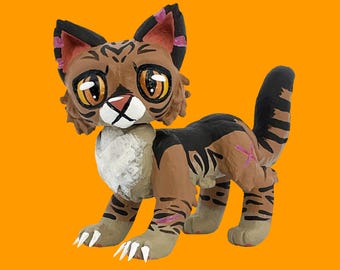 Könnte beinhalten: Handgefertigte Katzenfigur mit braunem und schwarzem Streifenmuster. Die Katze hat große, ausdrucksstarke Augen, eine weiße Schnauze und rosa Akzente an Ohren und Pfoten. Die Figur steht vor einem orangefarbenen Hintergrund.