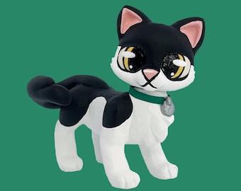 Könnte beinhalten: Keramik-Katzenfigur in stehender Pose, mit einem schwarz-weißen Tuxedo-Muster. Die Katze hat große, ausdrucksstarke Augen mit gelben und schwarzen Details, rosa Ohren und ein grünes Halsband mit einem silbernen Anhänger. Der Hintergrund ist einfarbig grün.