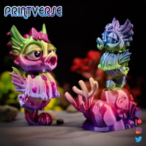 May include: Two vibrant 3D-printed sea creature figurines. One is a fish-like creature with an open mouth, and the other is a seahorse. The figurines are painted in pink, purple, green, and blue. The Printverse logo is visible.