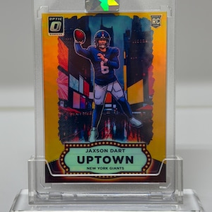 Puede incluir: Una tarjeta de fútbol Jaxson Dart Uptown New York Giants en una funda protectora. La tarjeta presenta a un jugador con uniforme azul lanzando un balón contra un fondo de paisaje urbano. La tarjeta es dorada con el logo Optic.