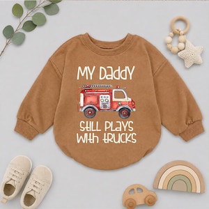 May include: A tan long-sleeve sweatshirt featuring a graphic of a red fire truck in the center. The text above the truck reads "MY DADDY" and the text below the truck reads "STILL PLAYS WITH TRUCKS" in a white, rounded, sans-serif font.