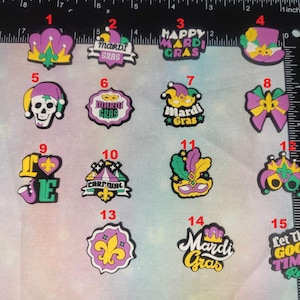 May include: A collection of colorful Mardi Gras-themed charms. Designs include crowns, masks, fleur-de-lis, and text like "Happy Mardi Gras" and "Let the Good Times Roll." The charms are made of a durable material and are suitable for various crafting projects.