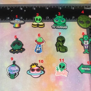 May include: A collection of colorful alien-themed charms. Designs include a UFO with a rainbow, green aliens with various expressions, and a glittery alien figure. Some charms have text that reads "ALLERGIC TO HUMANS" and "your favourite mama".