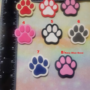 May include: A collection of eight colorful paw print-shaped embellishments. The paw prints feature various color combinations, including red, pink, black, white, and blue. The image includes a ruler for size reference. The text "Black Base" and "Navy Blue Base" is visible.