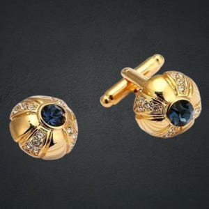 Könnte beinhalten: Ein Paar goldfarbene Manschettenknöpfe mit rundem Design. Jeder Manschettenknopf hat einen dunkelblauen Mittelstein, der von kleinen, klaren Kristallen umgeben ist. Die Manschettenknöpfe haben einen klassischen, eleganten Stil.